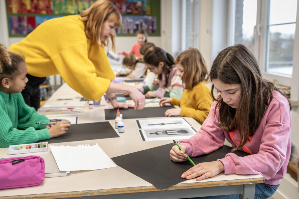 Beeldatelier Hooglede Enkele meisjes tekenen op een groot zwart blad papier onder begeleiding van de leerkracht.